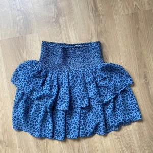 Blå leopardmönstrad kjol - Säljer en blå kortkjol med leopardmönster och volanger. Kjolen har en smockad midja som ger en bekväm passform. Perfekt för att ge en trendig touch till din outfit!