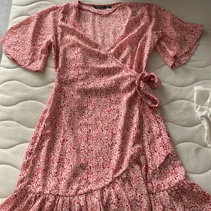 Röd blommig omlottklänning från Amelie & Me - Charmig röd omlottklänning med blommigt mönster från Amelie & Me. Klänningen har korta ärmar och en knytning i midjan för en smickrande passform. Perfekt för en somrig look! 🌸