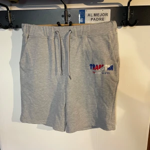 Grå shorts från Trapstar - Snygga grå shorts från Trapstar med broderad logga i rött, vitt och blått. De har en bekväm passform med snörning i midjan för justerbarhet. Perfekta för en avslappnad stil.