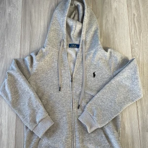 Grå hoodie från Ralph Lauren - Säljer en stilren grå hoodie från Ralph Lauren med dragkedja och klassisk logga på bröstet. Perfekt för en avslappnad look. Den har långa ärmar och en justerbar huva med snören. !OBS! (Det är en large men passar som en small)