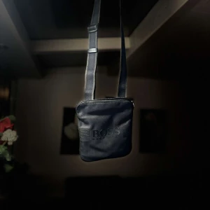 Svart axelväska från Boss - Snygg svart axelväska från Boss med justerbar rem. Väskan har en stilren design med Boss-logotypen på framsidan. Perfekt för att bära dina nödvändigheter med stil.