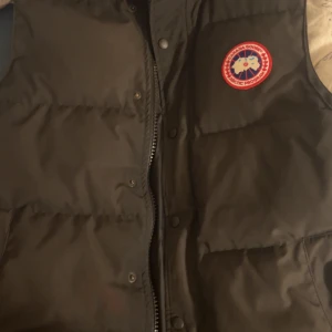 Svart dunväst från Canada Goose - Säljer en svart dunväst från Canada Goose med dragkedja och ett broderat märke på bröstet. Västen är perfekt för kyligare väder och har en stilren design. Köpt för 2 år sedan
