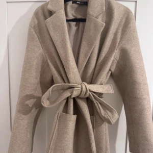 Beige lång kappa från Gina Tricot - Snygg beige lång kappa från Gina Tricot med knytbälte i midjan. Den har en klassisk krage och två stora fickor framtill. Perfekt för att hålla stilen under kyligare dagar.