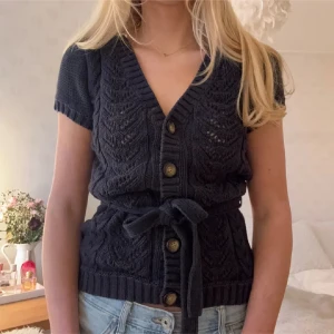 Mörkblå stickad kofta med knytband - Säljer en mörkblå stickad kofta med korta ärmar och knappar framtill. Koftan har ett fint mönster och ett knytband i midjan för en snygg passform. Perfekt för lager-på-lager-stil.