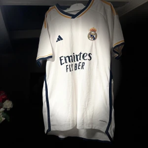 Vit Real Madrid fotbollströja 23/24 - Säljer en vit Real Madrid fotbollströja från Adidas med korta ärmar. Tröjan har klubbens emblem på bröstet och sponsorlogga framtill. Detaljer i guld och mörkblått ger en snygg kontrast. Perfekt för alla fans av laget!