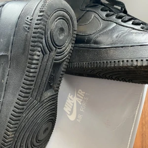 Svarta Nike Air Force 1 - Säljer ett par klassiska svarta Nike Air Force 1. Skorna har en hög modell med snörning och är tillverkade i skinn. Perfekta för en stilren look.