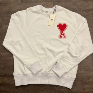 Vit sweatshirt från Ami - Säljer en stilren vit sweatshirt från Ami med ett rött hjärta och bokstaven A broderat på bröstet. Tröjan har långa ärmar och en rund halsringning. Perfekt för en avslappnad stil. Tillverkad av ekologisk bomull. Den är helt ny med inga slitningar.