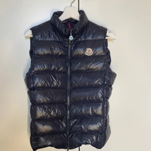 Moncler dunväst, navy - Dunväst från Moncler med dragkedja och logotyp på bröstet. Perfekt för kyligare dagar när du vill hålla stilen. Västen har en glansig finish och hög krage för extra värme.