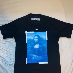 Mona Lisa Off-White tshirt - Snygg svart t-shirt från Off-White med ett stort blått Mona Lisa-tryck på framsidan och ikoniska vita pilar på baksidan. Perfekt för dig som vill ha en unik och stilren look. Passar bra till både jeans och sneakers.