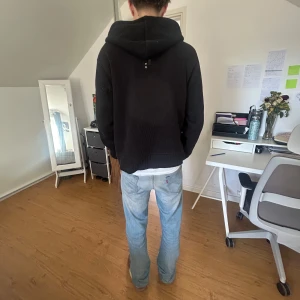 Svart hoodie med dragkedja - Säljer en svart hoodie med dragkedja och huva. Tröjan har ribbade muddar och två fickor framtill. Perfekt för en avslappnad stil.