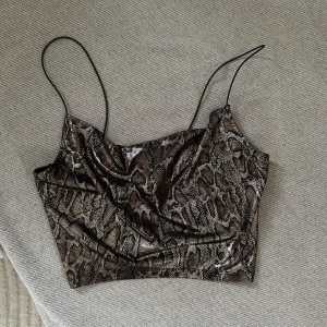 Ormskinnsmönstrad topp med tunna axelband - Trendig ormskinnsmönstrad topp med tunna axelband. Perfekt för en kväll ute eller en stilfull vardag. Toppen har en glansig finish som ger en extra touch av glamour.