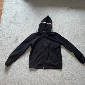 Svart vindjacka från Moncler  - Säljer en svart vindjacka från Moncler med huva och dragkedja. Jackan har en vit och röd rand på huvan, två fickor med knappar och justerbar dragsko. Klassisk Moncler-logga på bröstet och detaljer i metall.