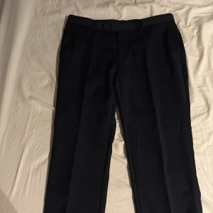 Mörkblå kostymbyxor från Dressmann - Säljer ett par mörkblå kostymbyxor från Dressmann i slim fit-modell. Byxorna har raka ben, bälteshällor och knäppning fram samt bakfickor med knapp. Perfekta för en stilren look. Byxorna är i storlek 32/32 och passar dej som är runt 180. Använda cirka 3 gånger. Hör av er vid frågor och funderingar