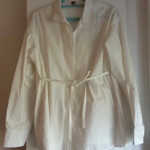 Vit skjorta med lång ärm och knytbälte i midjan - Säljer en stilren vit skjorta med lång ärm och knytbälte i midjan. Skjortan har klassisk krage och knäppning framtill, perfekt för en clean och enkel look.