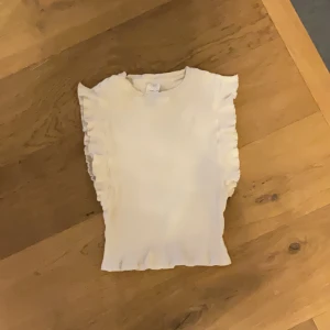 Vit ribbad topp med volang från Zara - Säljer en söt vit ribbad topp från Zara i storlek 140 (9-10 år). Toppen har volangdetaljer längs axlarna och är ärmlös. Perfekt för vår och sommar!