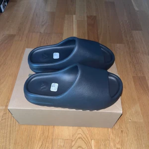 Svarta Yeezy Slide sandaler - Säljer ett par svarta Yeezy Slides helt nya. Jag råka klippa StockX taggen innan jag kolla om det passar mig så nu måste jag sälja dem. Perfekt för sommaren och super skönna. Det är storlek 43 men passar också 42