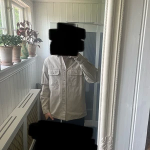 Beige overshirt från H&M - Säljer en stilren beige overshirt från H&M med två bröstfickor och knappar framtill. Skjortan har klassisk krage och lång ärm, perfekt att bära öppen eller stängd över en t-shirt.