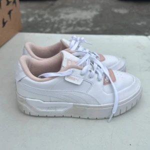 Vita sneakers från Puma med ljusrosa detaljer - Säljer ett par fräscha vita Puma sneakers med ljusrosa detaljer och klassisk snörning. Skorna har en chunky sula och är perfekta för dig som gillar en sportig och stilren look. Puma-logga på sidan och plös.