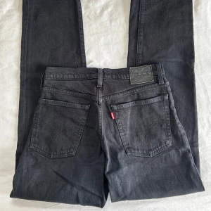 Svarta Levi's Wedgie Straight jeans - Säljer ett par svarta Levi's Wedgie Straight jeans med klassisk femficksdesign och knappar i gylfen. Jeansen har raka ben samt detaljer i silver och koppar. Perfekta för dig som gillar en tidlös look. Byxorna är W26 (ca 36)
