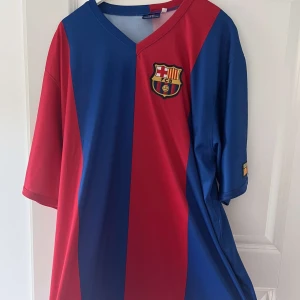 FC Barcelona fotbollströja Ronaldinho 10 retro  - Säljer en klassisk FC Barcelona fotbollströja med Ronaldinho och nummer 10 på ryggen. Tröjan är kortärmad med breda vertikala rödblå ränder, klubbmärke på bröstet och tryck i guld på ryggen. Perfekt för dig som älskar fotboll och legendariska spelare.