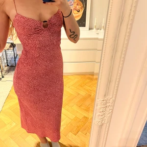midiklänning med ringdetalj - Säljer en röd/rosa/vit mönstrad klänning med smala axelband och snygg ringdetalj framtill. Klänningen har figurnära passform och är perfekt för sommaren. Mönstret är smått och ger en livlig känsla.
