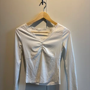 Vit långärmad topp med spetsdetalj - Säljer en vit långärmad topp med v-ringning och spetsdetalj vid halsen. Toppen har en lätt rynkning framtill och är tillverkad i ett mjukt, ribbat material. Perfekt att styla till jeans eller kjol.