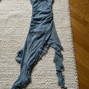 Blå klänning med volang - Säljer en blå klänning från Zara i storlek S. Klänningen har volangdetaljer längs kanten och är tillverkad i ett plisserat, lätt material. Asymmetrisk nederkant och smickrande passform gör den perfekt för sommarens tillfällen.