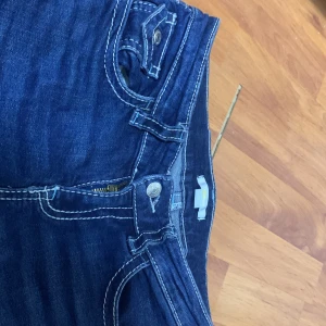 Mörkblå jeansbyxor med kontrastsömmar - Säljer ett par mörkblå jeansshorts med tydliga vita kontrastsömmar och klassiska fickor fram och bak. Byxorna har knapp- och dragkedjestängning samt dekorativa detaljer på bakfickorna. Perfekta för en avslappnad stil.