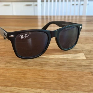Svarta solglasögon från Ray-Ban - Klassiska svarta solglasögon från Ray-Ban med Wayfarer-form. Bågarna är mattsvarta och har silvriga detaljer vid fronten. 