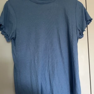 Blå t-shirt/topp med volangärm - Säljer en ljusblå ribbad t-shirt med korta ärmar och volangdetaljer vid ärmslut och nederkant. Perfekt till vardags och enkel att matcha med jeans eller kjol. Säljer samma på vinted så kontakta gärna innan ni trycker på ”köp”!