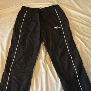 Svarta träningsbyxor från FSBN med vita detaljer - Svarta träningsbyxor från FSBN med vita ränder längs benen och dragkedjor vid fickorna. Byxorna har resår i midjan med snörning och är perfekta för sport eller chill. Märke tryckt på ena benet.