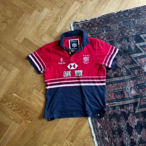 Piké Hong Kong rugby - Säljer en klassisk rugbytröja från Kukri i rött och marinblått med broderade detaljer och tryck från Hong Kong Rugby. Tröjan har krage, knappar och vita ränder över bröstet och ärmarna. Perfekt för dig som gillar sportig stil!