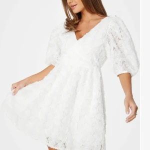 Vit blommig studentklänning med puffärm - Superfin vit kortklänning med v-ringning och omlottkänsla. Klänningen har 3/4-puffärmar och är täckt av blommiga 3D-detaljer som ger en romantisk look. Perfekt för studenten!