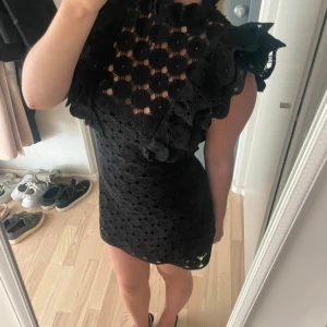 Svart spetsklänning med volang - Säljer en svart kort klänning i virkat och volangdetaljer vid axlarna. Helt slutsåld på H&M. Klänningen är ärmlös och har hög hals. Materialet känns lyxigt.