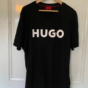Hugo Boss tröja - Säljer en svart t-shirt från Hugo Boss med stor vit logotyp på bröstet. Klassisk rund halsringning och korta ärmar. Perfekt för en stilren och enkel look. Köpt för 600kr. Lite liten i storleken, står XL, men skulle säga att den passar L bäst💯knappt använd 