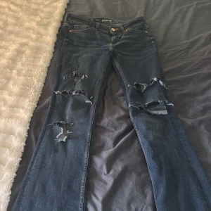 Mörkblå bootcut jeans med slitningar - Mörkblå bootcut jeans från Denim med flera slitningar och hål på både fram- och baksida av benen. Klassisk femficksmodell med knapp och dragkedja. Perfekta för en avslappnad och trendig look.