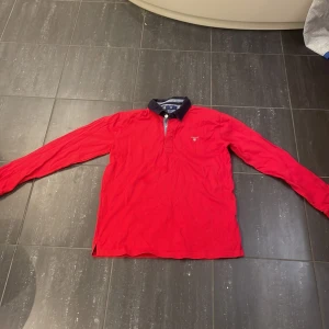 Röd långärmad pikétröja från Gant - Säljer en klassisk röd långärmad pikétröja från Gant med mörkblå krage och knappslå. Tröjan har en liten broderad logga på bröstet och passar perfekt till en avslappnad men stilren look.