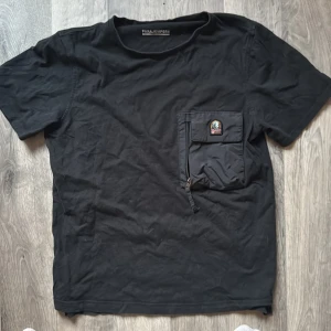 Svart t-shirt från Parajumpers med ficka - Säljer en svart t-shirt från Parajumpers med en stor ficka med dragkedja och logga på bröstet. T-shirten har normal passform och är kortärmad, perfekt för en stilren och avslappnad look. Oklar storlek men passar mig som är 180 och passade mig när jag var ish 165