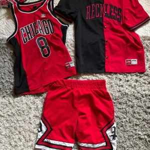 Röd och svart basketinspirerad t-shirt från FSBN - Säljer en röd och svart t-shirt från FSBN med texten 'RECKLESS' framtill. T-shirten har korta ärmar, knappar framtill och sportig design med kontrastfärgade detaljer. Perfekt för dig som gillar streetwear och sportig stil.