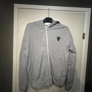 Grå hoodie med dragkedja från Comme des Garçons Play - Säljer en grå hoodie från Comme des Garçons Play med dragkedja och huva. Tröjan har det klassiska svarta hjärta-logotypen broderat på bröstet och två fickor framtill. Perfekt för en avslappnad stil. Litet hål på armen, man märker inte av det. Passar small. Passar inte priset så kom med bud!