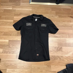 Svart kortärmad skjorta från Dickies - Säljer en svart kortärmad skjorta från Dickies med broderade patches på bröstet. Skjortan har klassisk krage, knappar framtill och två bröstfickor med lock. Perfekt för en avslappnad och cool stil.