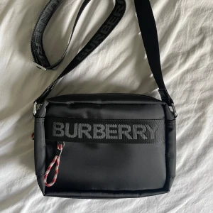 Svart axelremsväska  - Säljer en helt oanvänd svart axelremsväska. Dan har många fack och får platts med mycket. Väskan har loggor på många olika ställen. Priset kan diskuteras.