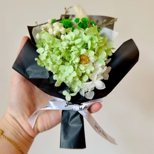 torkade blommor - en liten bukett med gröna och vita torkade blommor, inslagna i svart papper och dekorerad med ett vitt band. Buketten har en kompakt och rund form.