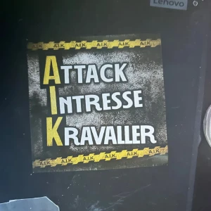 AIK Attack Intresse Kravaller affisch - Affisch med texten 'AIK Attack Intresse Kravaller' i svart, vitt och gult. AIK-logotypen och färgerna är tydliga, med gul text på svart bakgrund och AIK-tejp runt kanterna. Perfekt för AIK-supportern som vill visa sitt engagemang. 11st