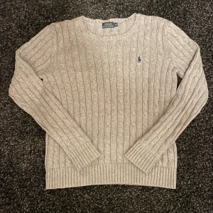 Grå kabelstickad tröja från Polo Ralph Lauren - Säljer en klassisk grå kabelstickad tröja från Polo Ralph Lauren med rund halsringning och broderad logga på bröstet. Tröjan har långa ärmar och ribbade muddar vid ärmslut och nederkant. Perfekt för lager-på-lager under kyliga dagar.