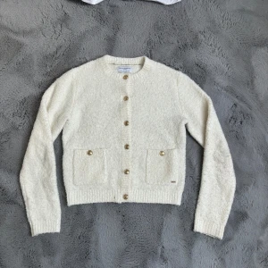 Vit stickad kofta  - Mysig vit stickad kofta med rund halsringning och guldknappar framtill. Två fickor framtill och ribbade muddar vid ärmslut och nederkant. Perfekt att slänga på sig när det är kyligt ute.