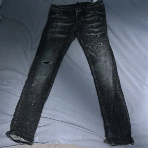 Svarta Dsquared2 jeans med slitningar - Säljer ett par svarta Dsquared2 jeans med coola slitningar och distressed detaljer. Jeansen har en klassisk femficksmodell och tydliga slitna partier framtill. Märkespatch bak i midjan. Perfekta för dig som gillar en edgy stil.