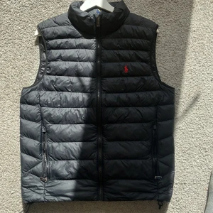 Svart dunväst från Polo Ralph Lauren - Säljer en svart dunväst från Polo Ralph Lauren med dragkedja framtill och två fickor med dragkedja. Västen har en diskret röd logga broderad på bröstet och quiltad design. Perfekt för lager-på-lager under kyliga dagar. Har digitalt kvitto, köpt på zalando för 3595kr och säljer min för endas 2400kr. vid snabb affär kan du få bättre pris, bara höra av sig vid minsta fundering ☺️har digitalt kvitto.