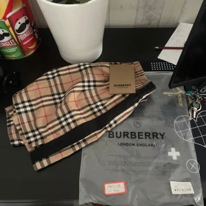 Rutig shorts från Burberry - Snygga shorts från Burberry i klassiskt beige rutigt mönster med svarta och röda linjer. Modellen har resår i midjan och snörning för extra komfort. Perfekta för sommaren och ger en lyxig känsla till din outfit.