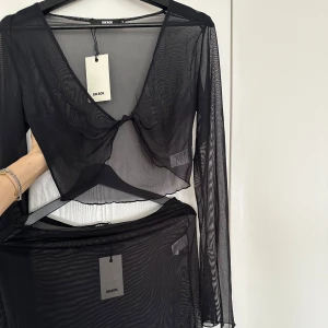 Beach cover-up set - Set med matchande topp och kjol. Fungerar som beach cover-up. Svart långärmad mesh topp med knut framtill i stl S. Svart mesh kjol i stl S. Nya, aldrig använda, med lappar kvar.